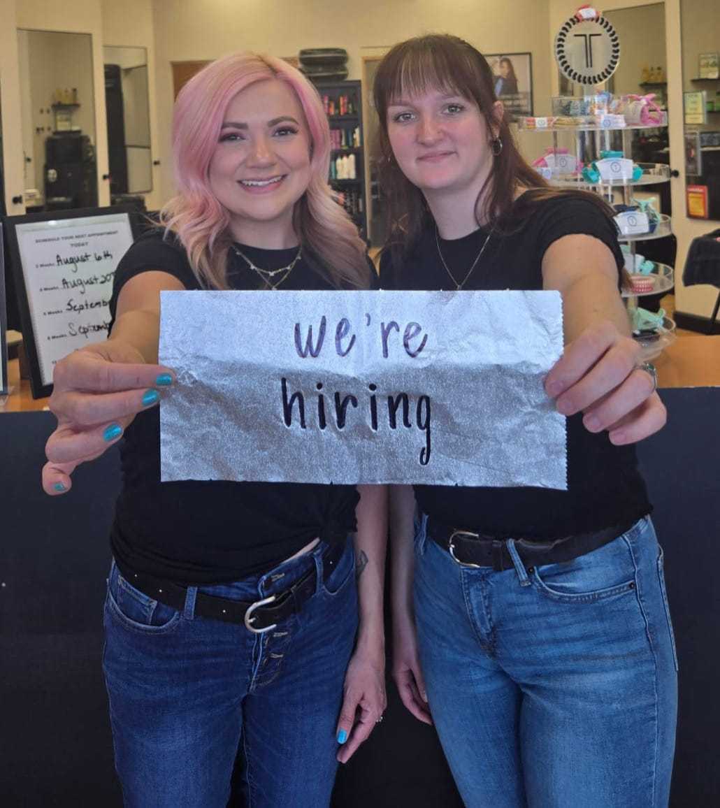 Two smiling women holding a "we're hiring" sign in a shop setting.